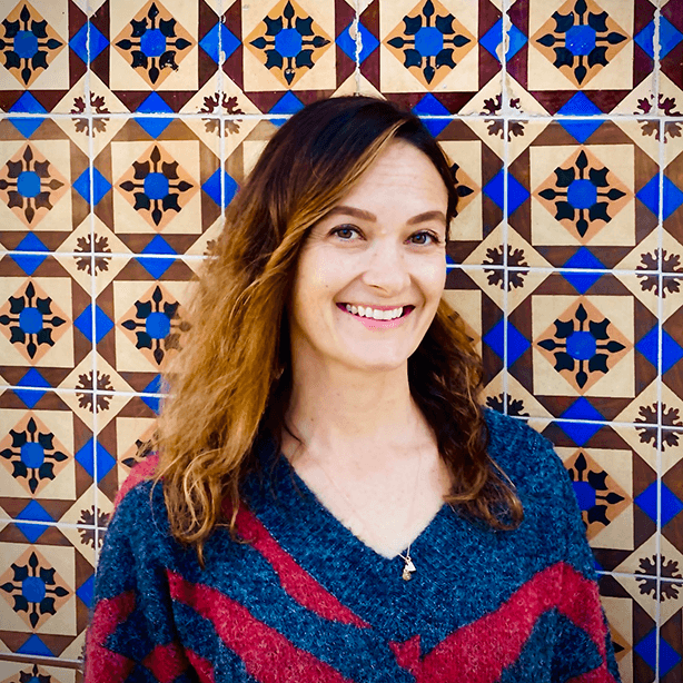 Woman with wavy hair wearing a blue and red sweater, smiling in front of a colorful geometric patterned tiled wall.