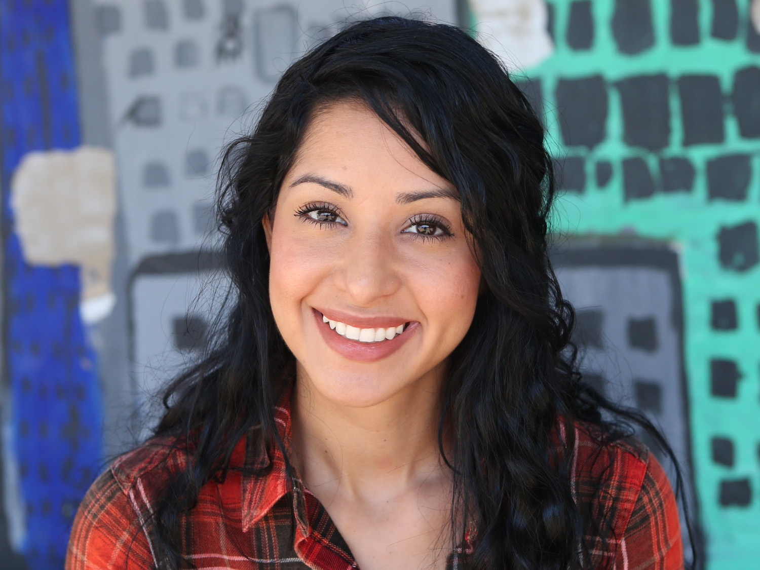 Smiling woman with long black hair, wearing a red plaid shirt, standing against a vibrant, abstract mural backdrop.