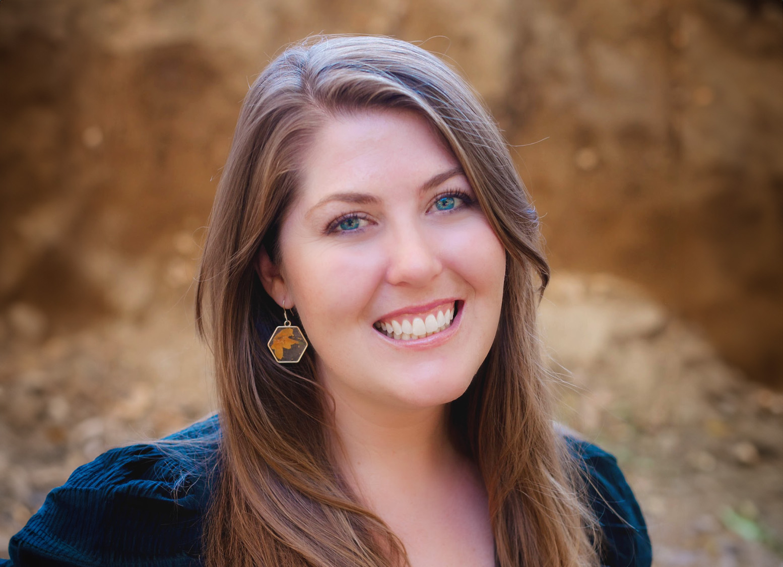 Smiling woman with long brown hair wearing a black top and geometric earrings, set against a blurred earthy background.