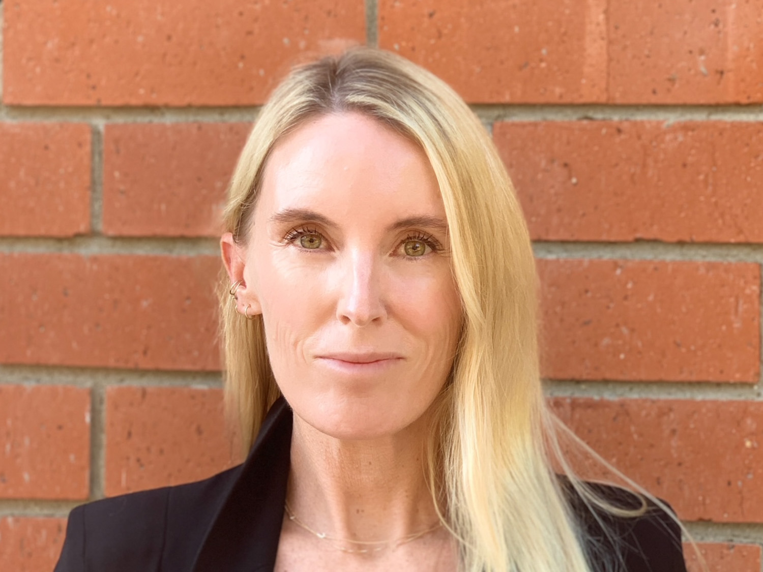 Woman with long blonde hair, wearing a black blazer, standing in front of a brick wall.