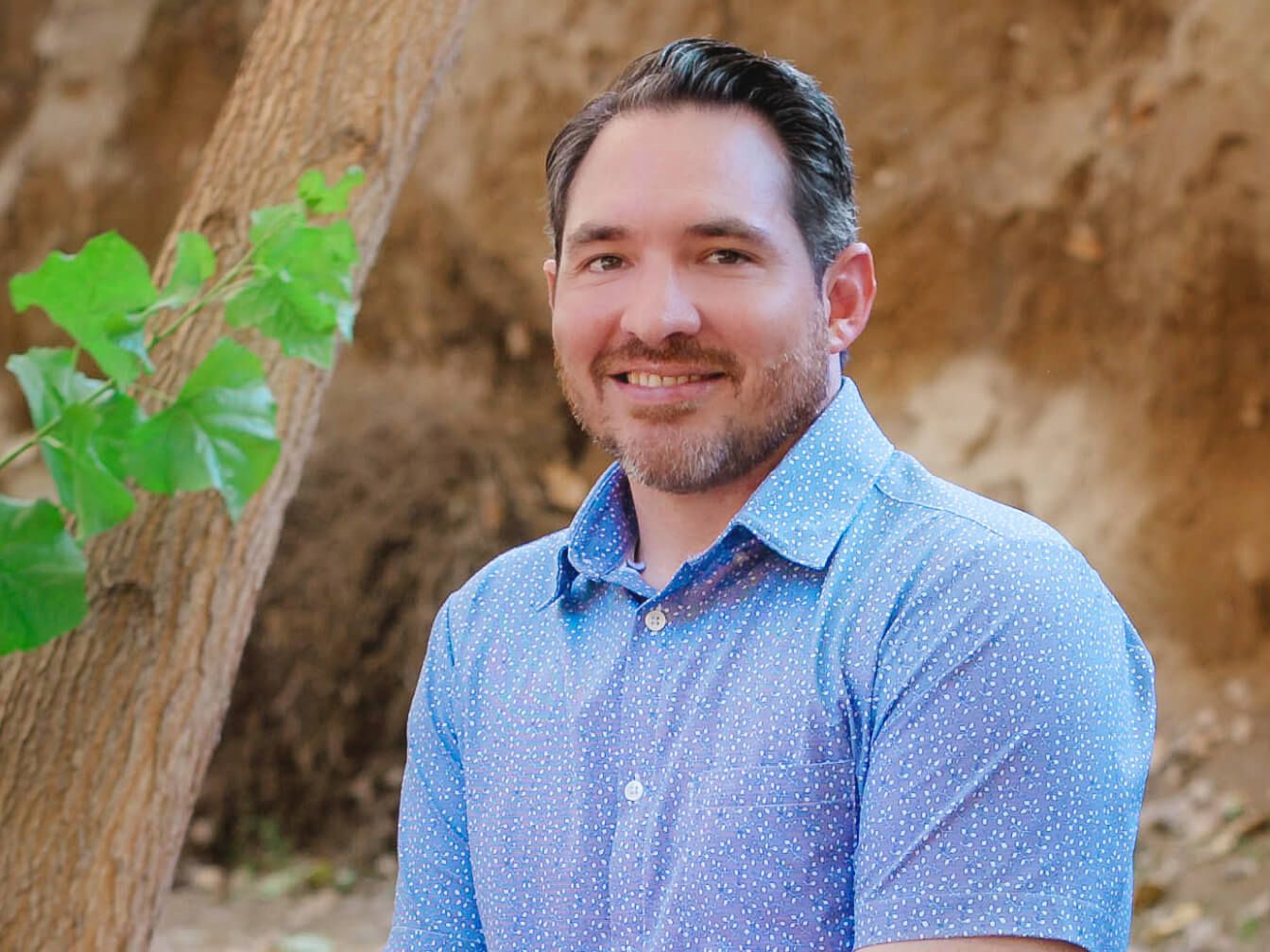 A person with short dark hair and a beard, wearing a light blue patterned shirt, is smiling while standing outdoors near a tree trunk and green leaves, with a blurred brown background.