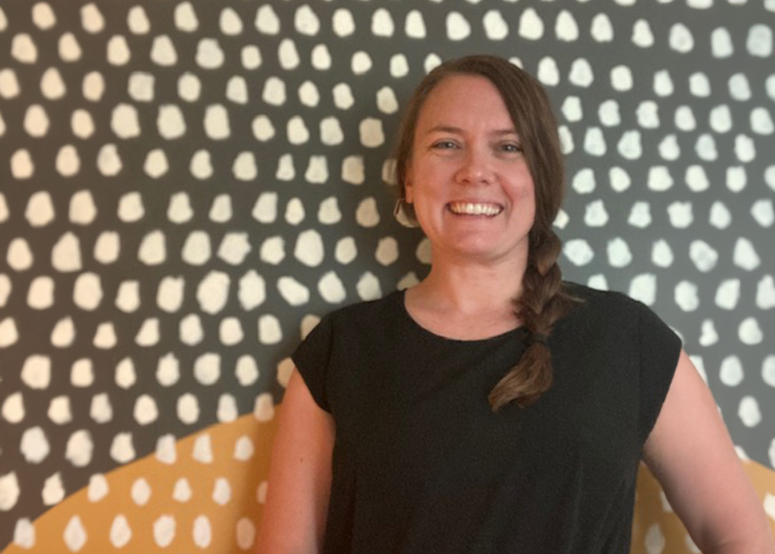 Smiling woman with a side braid wearing a black top standing in front of a patterned wall.
