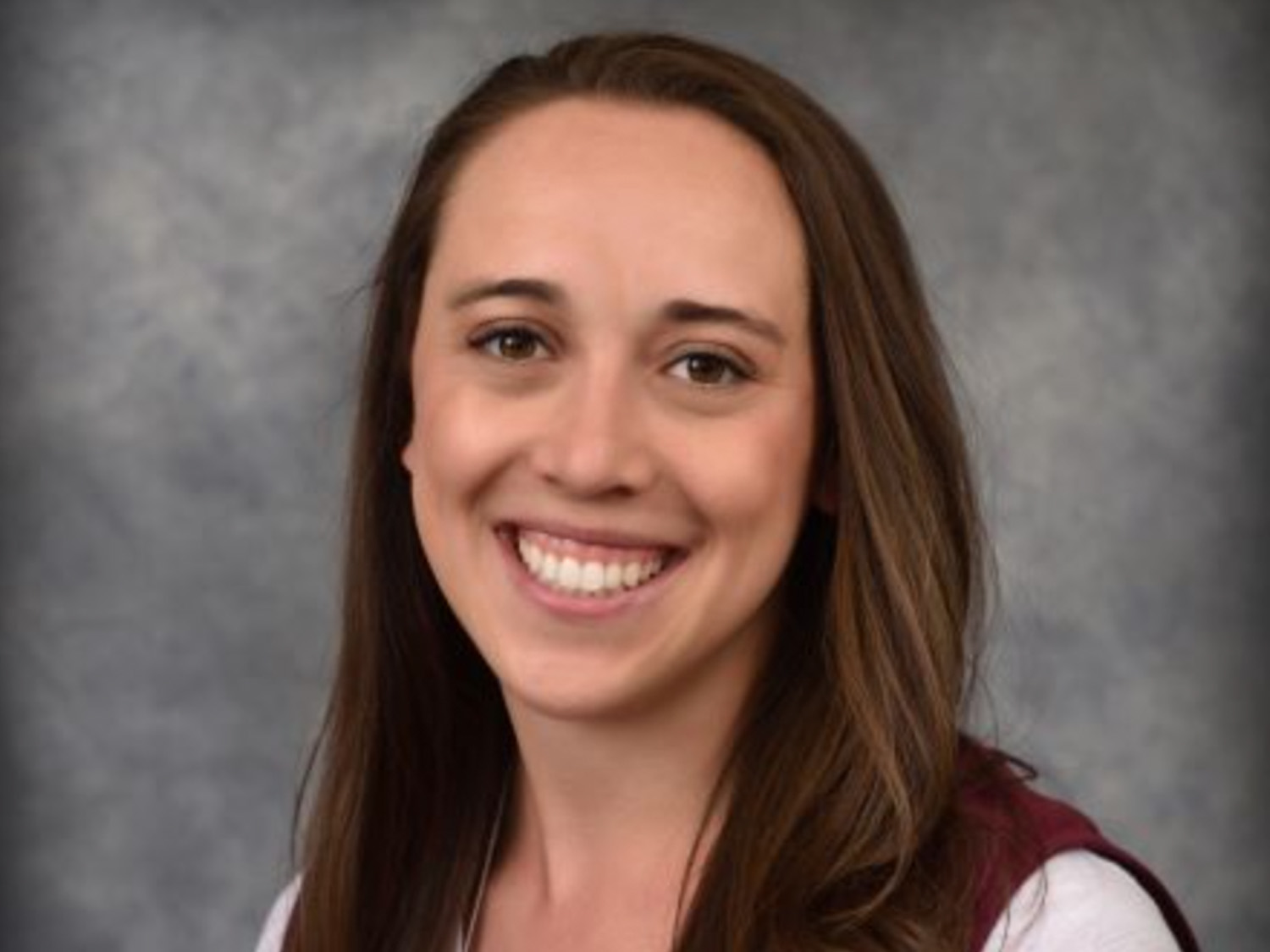 Smiling woman with long brown hair, wearing a white top and maroon vest, against a gray textured background.