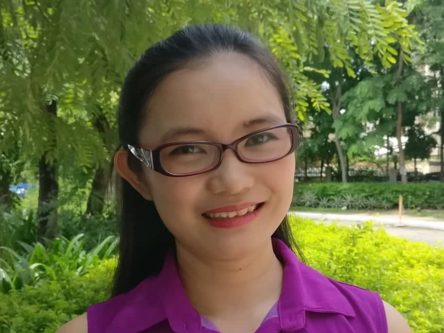 Young woman with dark hair wearing glasses and a purple shirt, smiling in an outdoor setting with lush green foliage in the background.