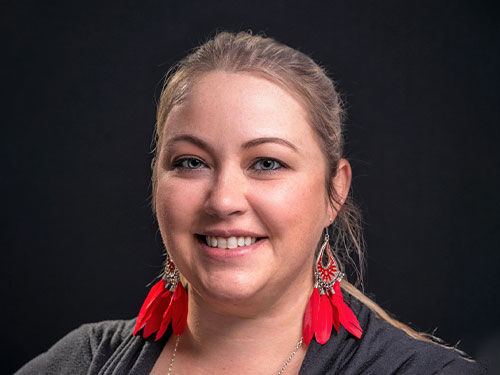 Smiling woman with long blonde hair, wearing bright red feather earrings and a dark top, against a black background.