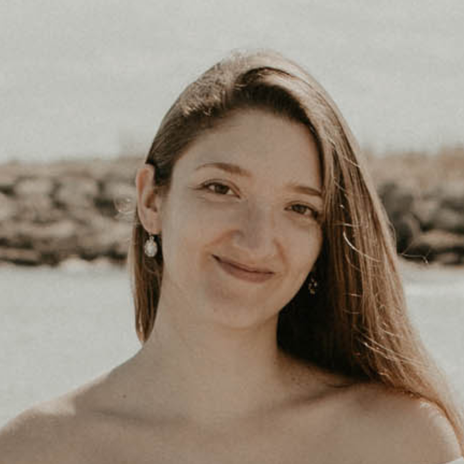 A young woman with long brown hair smiles warmly; she is outdoors with a blurred rocky background.