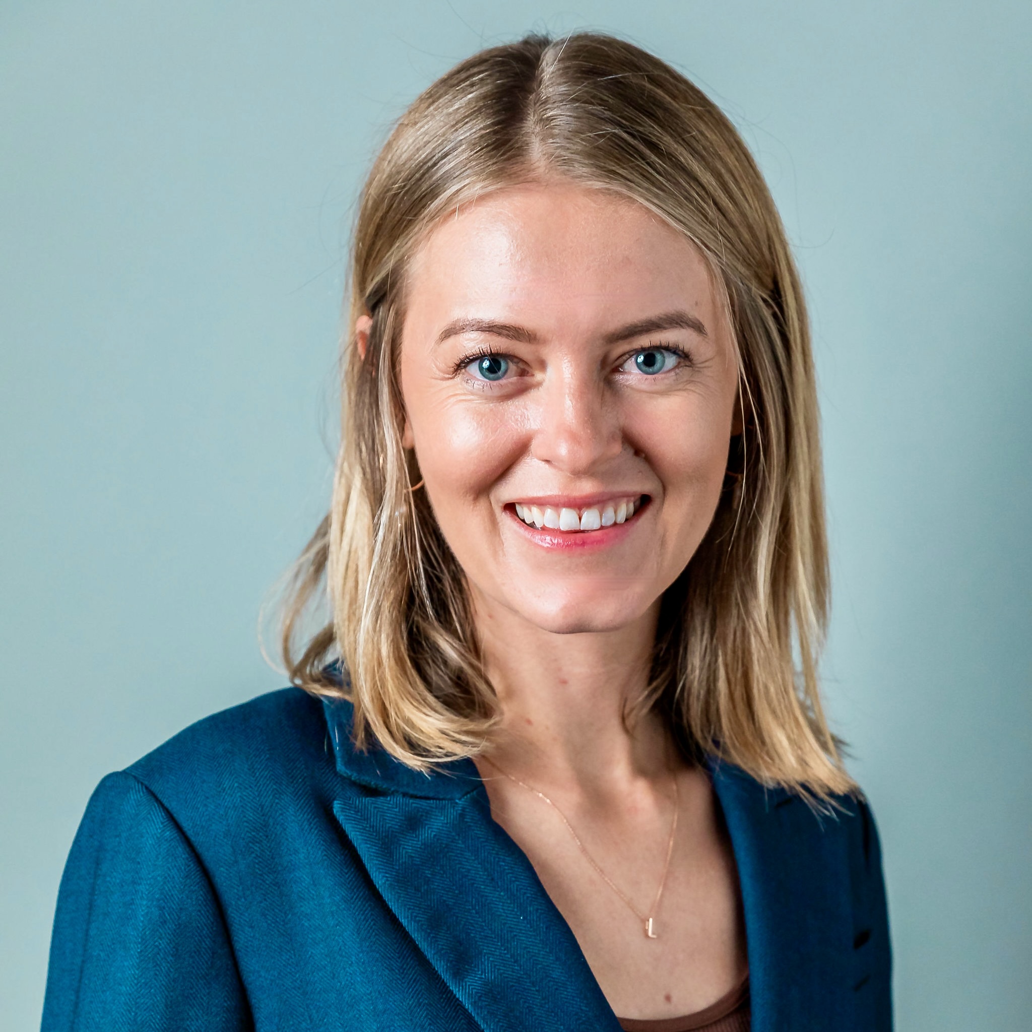 A woman with shoulder-length blonde hair and blue eyes, wearing a teal blazer over a dark top, smiling against a neutral background.