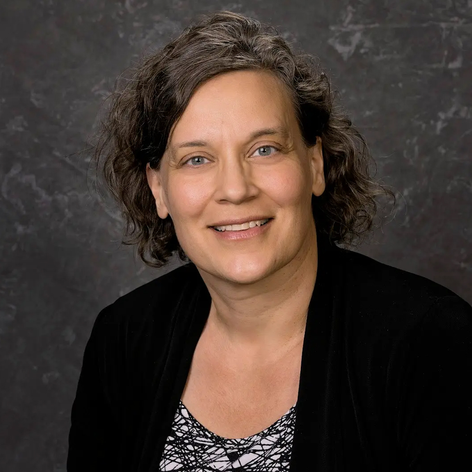 Woman with curly brown hair, wearing a black top over a patterned blouse, smiling in front of a dark gray background.