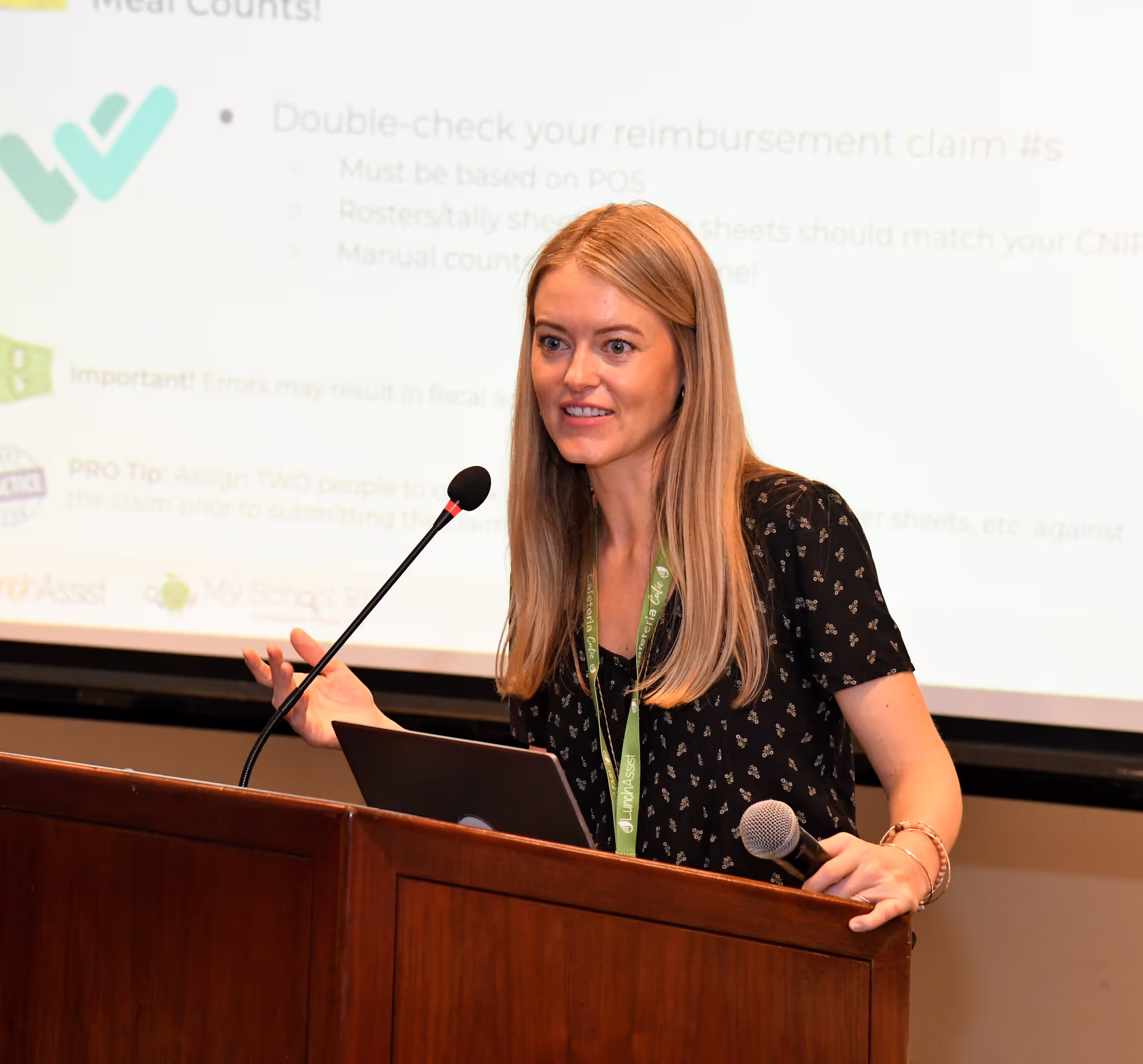 A woman with long blonde hair speaks into a microphone at a podium, holding a tablet, with a presentation slide projected in the background.