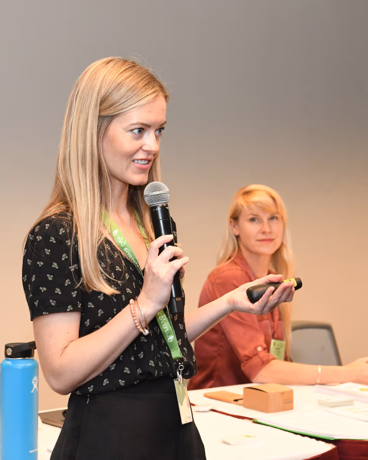 Two women in a conference room setting, one standing and speaking into a microphone while holding a remote, the other seated and looking on. They are at a table with papers and a blue water bottle. Both have long blonde hair and are wearing ID lanyards.