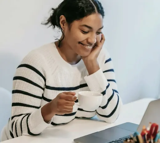 A person wearing a striped sweater sits at a table with a laptop, smiling and holding a white mug.