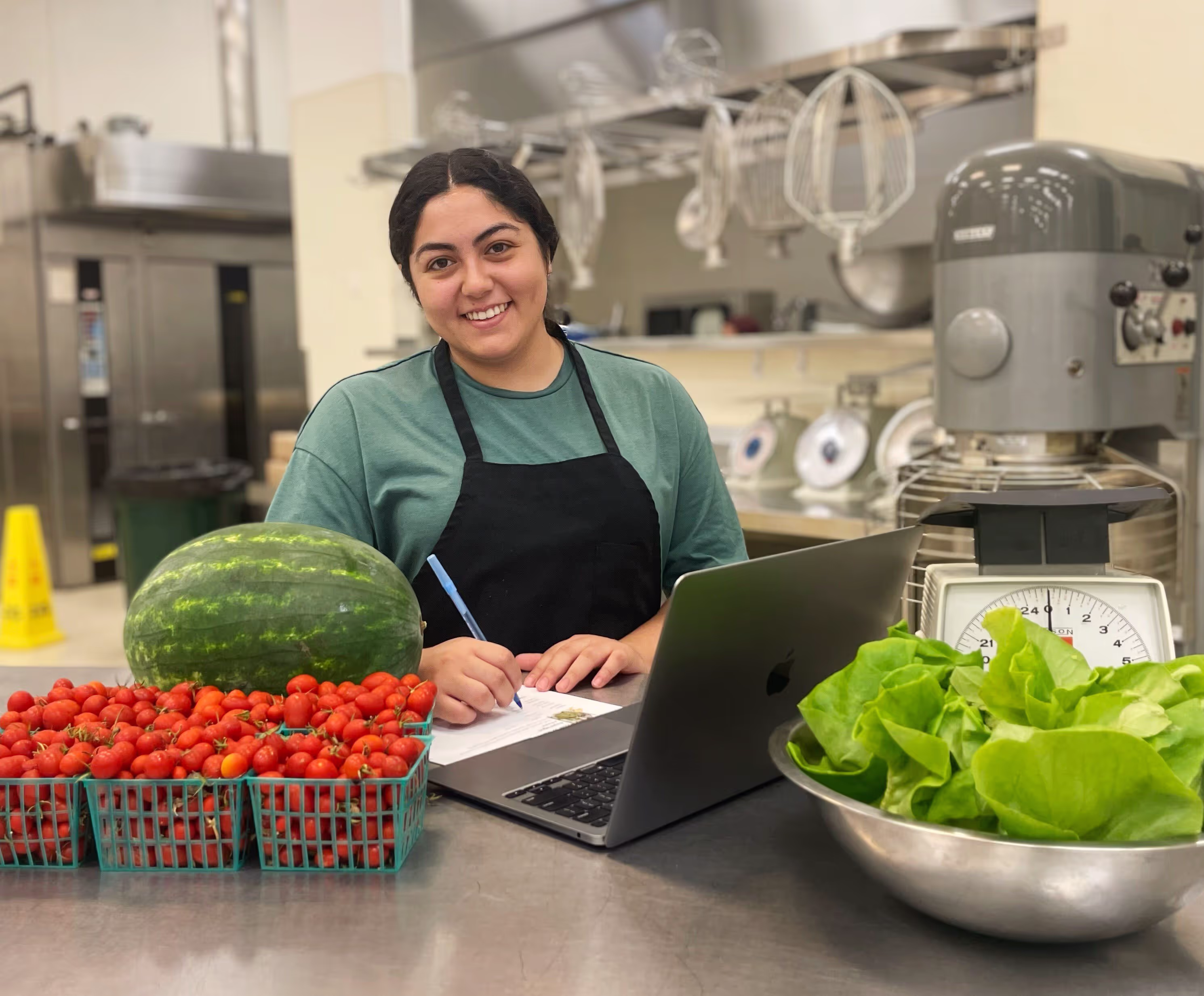 Person wearing a green shirt and black apron, standing in a kitchen with a laptop, notepad, and pen. In front of them are trays of cherry tomatoes, a watermelon, and a bowl of lettuce. Kitchen equipment and mixers are visible in the background.