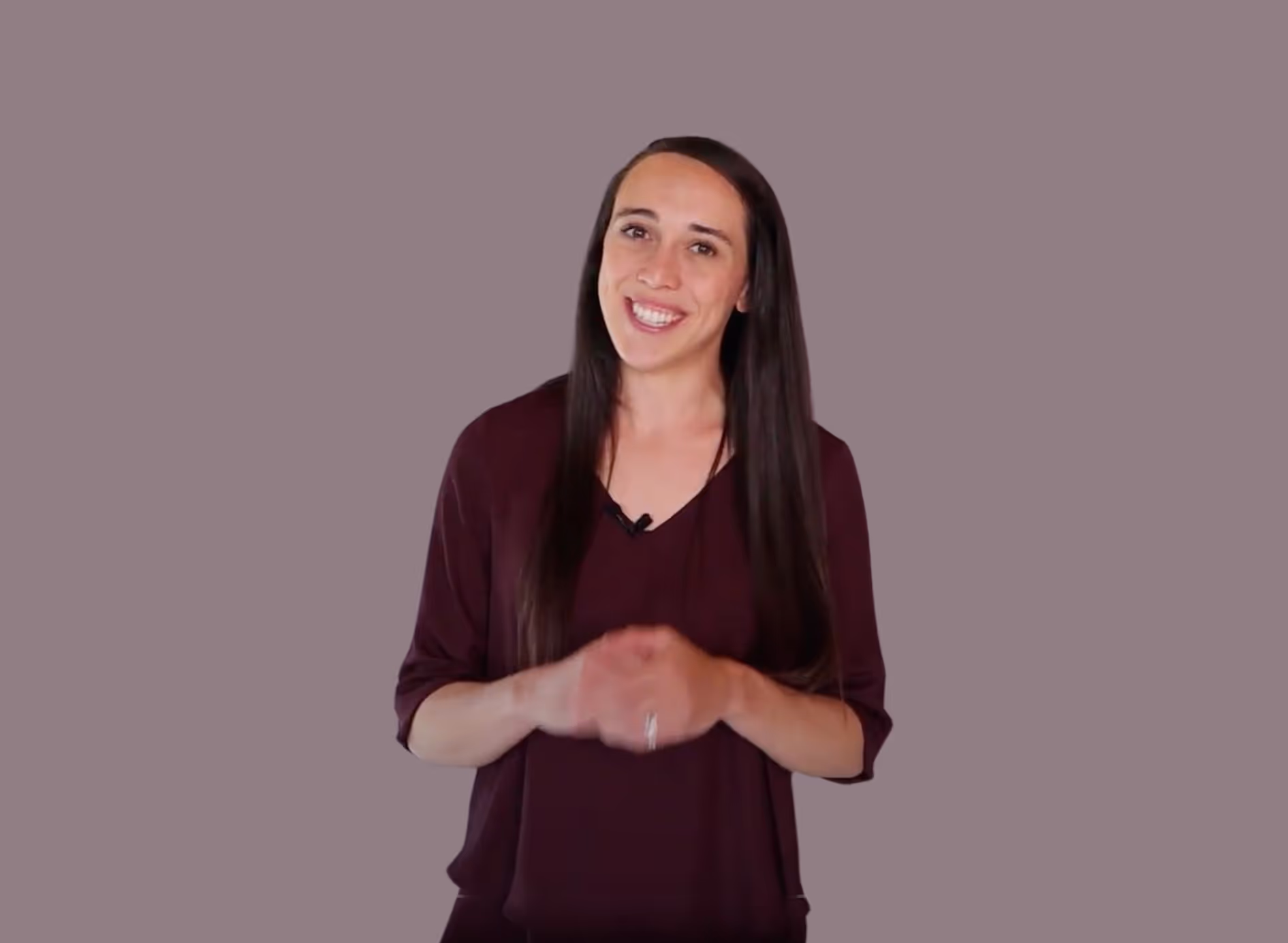 Smiling person with long dark hair, wearing a dark maroon top, standing in front of a plain gray background.