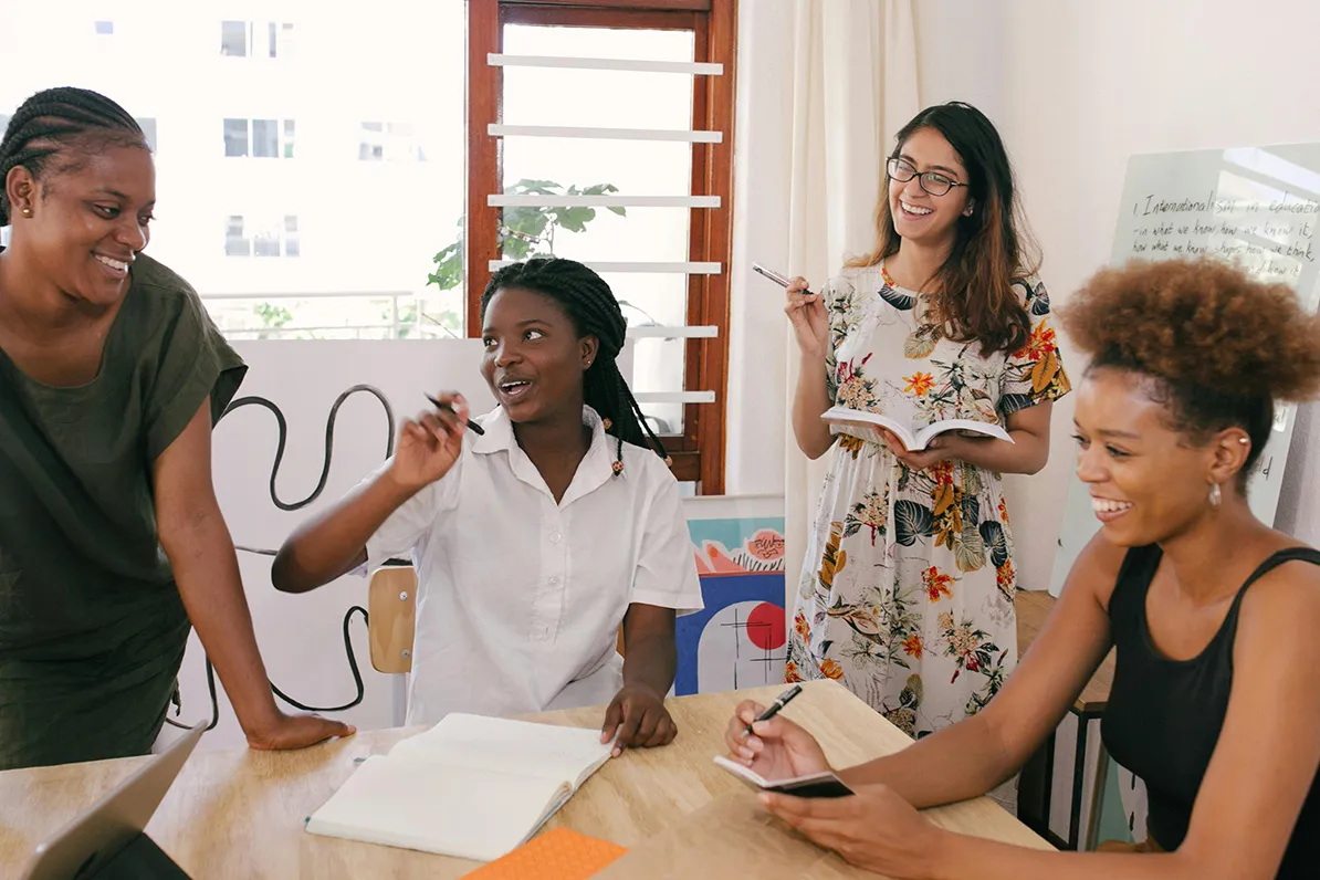 Four women in casual and business attire engage in a lively discussion around a wooden table, with notepads and papers, in a bright room featuring abstract art and a large window.