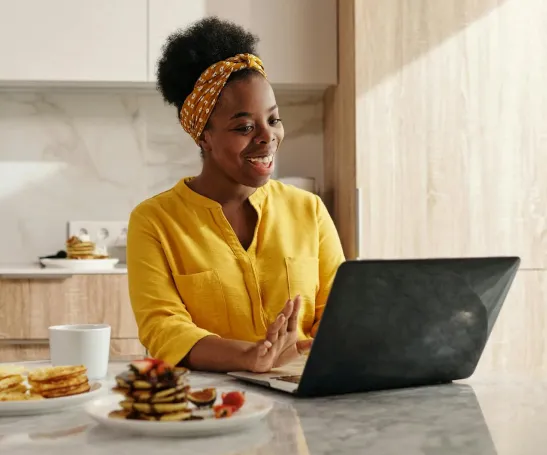 Smiling person wearing a yellow shirt and headband, sitting at a kitchen counter with a laptop, waving. A stack of pancakes with strawberries is in the foreground, and a white mug is nearby. Bright kitchen setting.