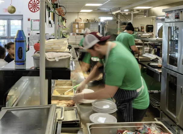 Person in a commercial kitchen wearing a green shirt and cap, preparing food with dishes in the foreground; kitchen equipment and supplies are organized in the background.