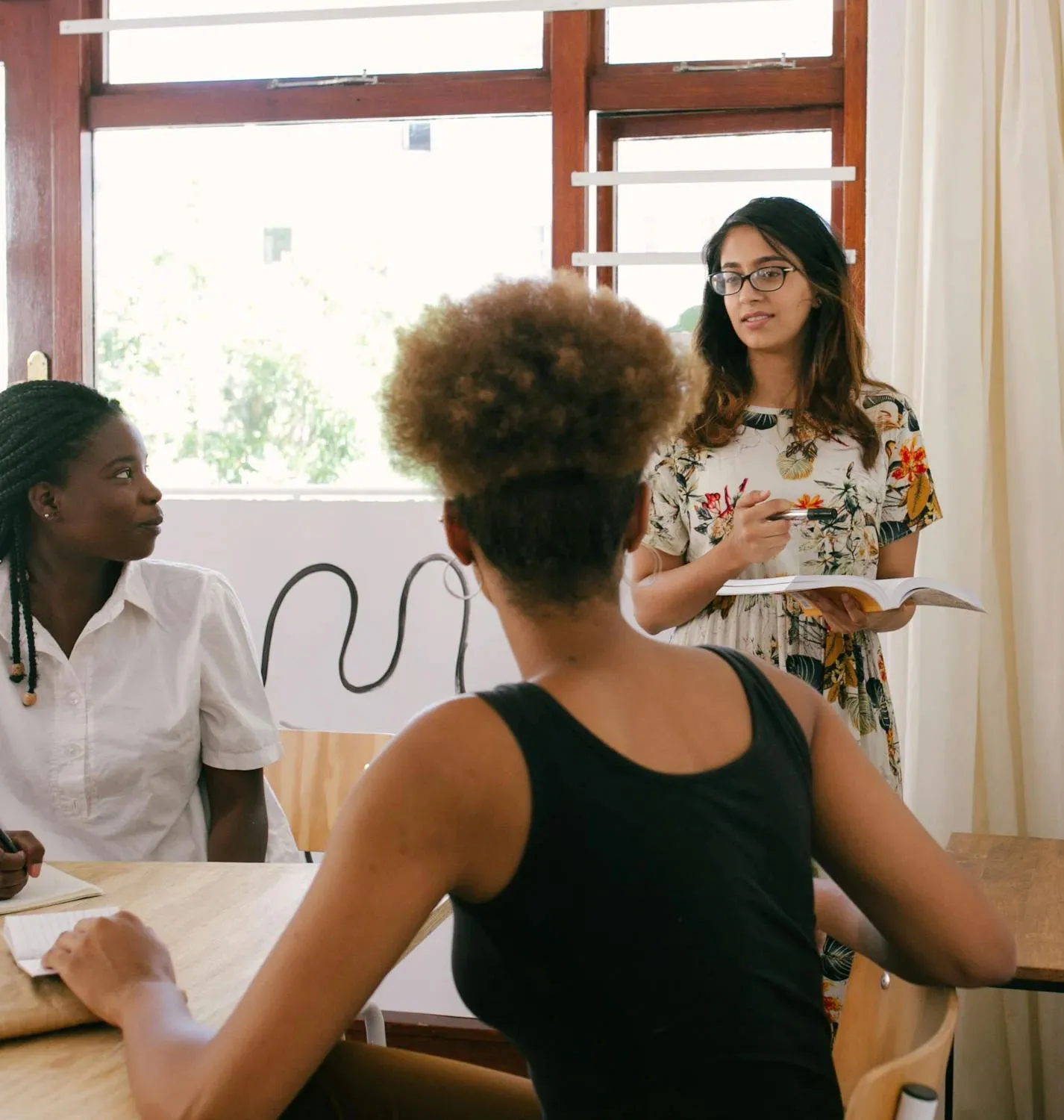 Three women engaged in a meeting; one standing and speaking with notes, wearing glasses and a floral dress, while the other two sit and listen intently at a table.