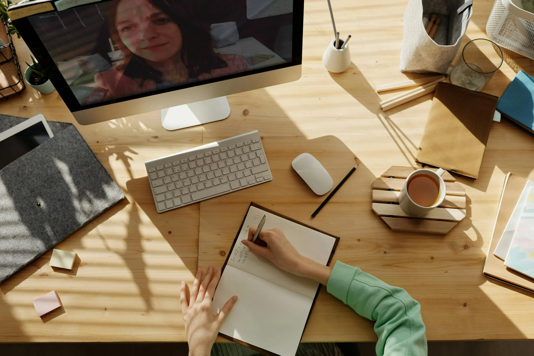 Person writing in a notebook at a wooden desk with a keyboard, mouse, and a mug beside them, sunlight casting shadows; a computer screen shows a video call with another person.
